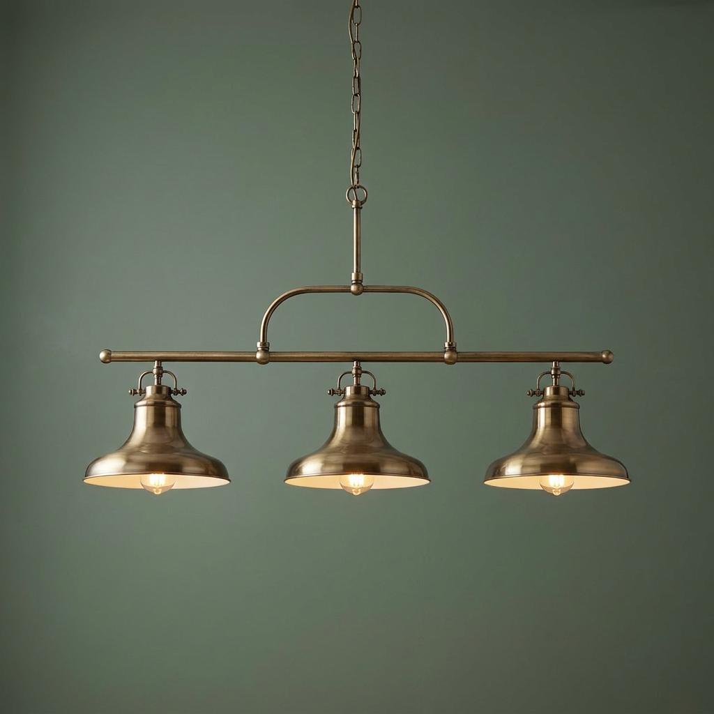 Three-light brass bar pendant with bell-shaped shades hanging against a green wall, emitting a warm glow.