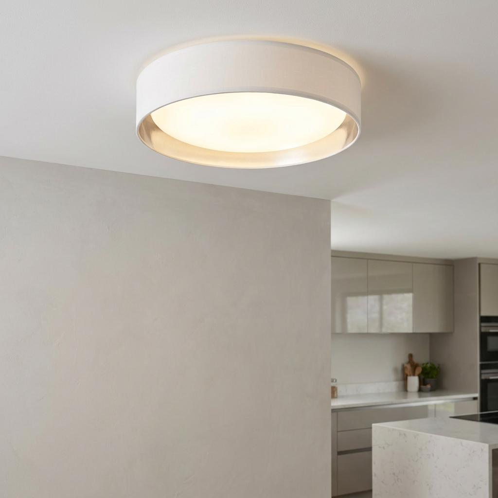 Modern flush ceiling light with a white fabric drum shade and silver interior lining, illuminated in a kitchen setting.