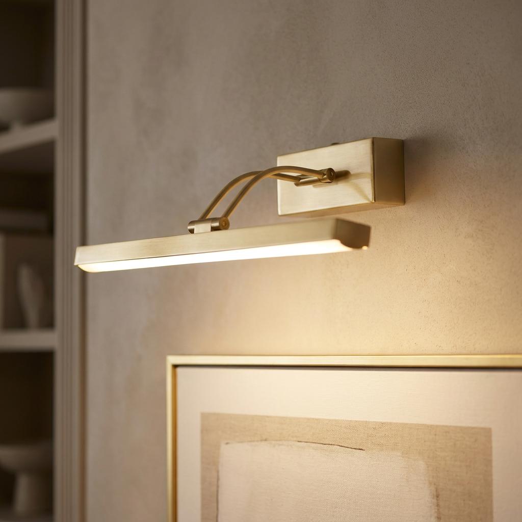 Brushed brass LED picture light mounted on a wall above a framed painting, providing a warm downward glow.