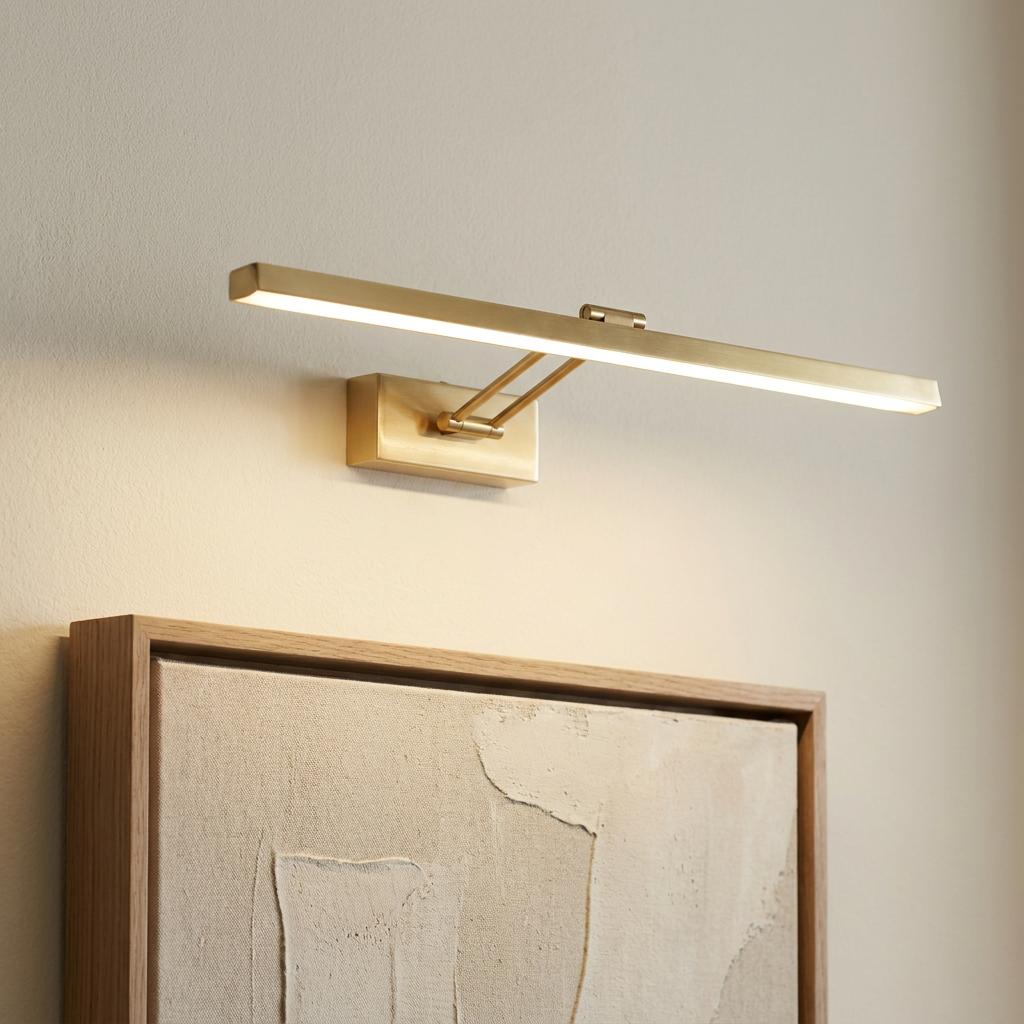 A modern brass LED picture light illuminating a textured piece of artwork on a neutral wall.