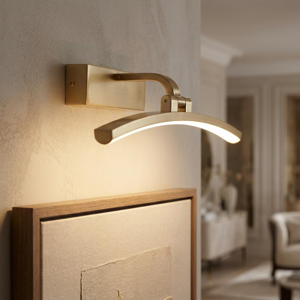 Sleek curved brass LED picture light mounted on a wall, illuminating a framed artwork in a modern interior.