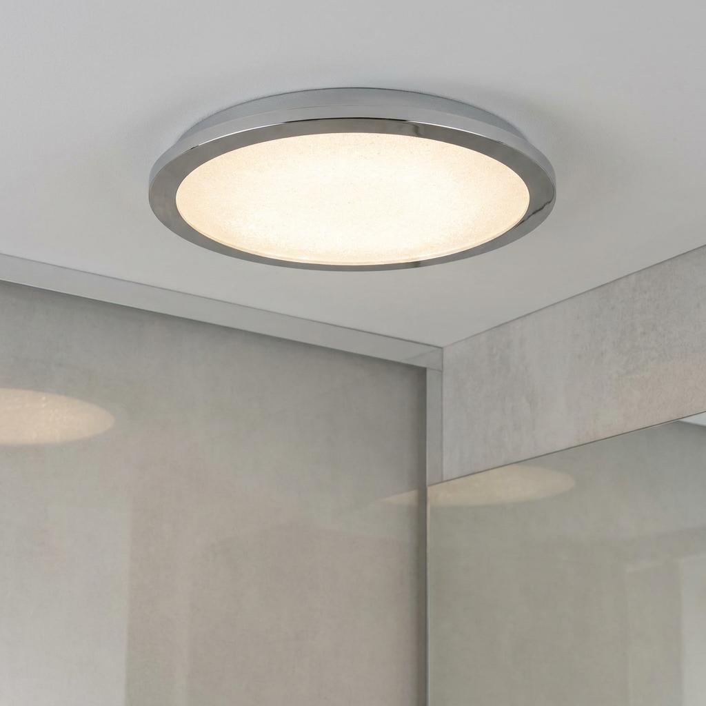 Circular chrome LED flush mount light with a crystal sand diffuser glowing on a white ceiling in a bathroom.