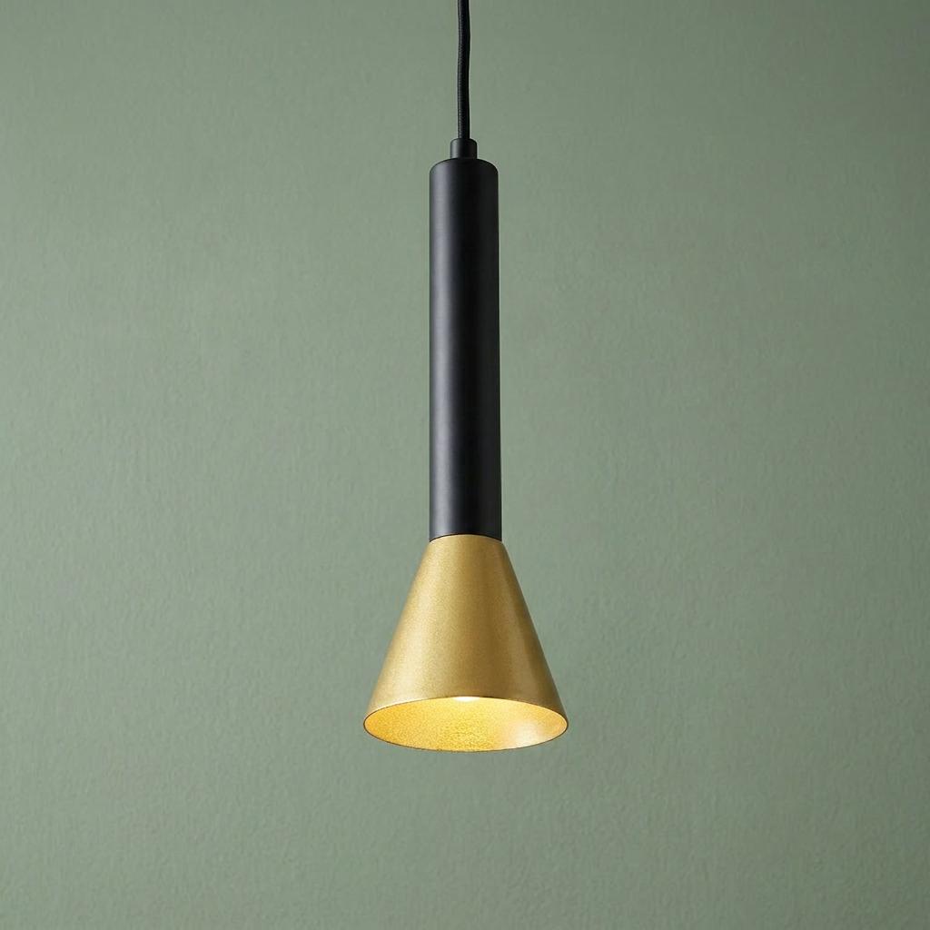 Modern black and gold pendant light with a conical shade, lit and hanging against a sage green wall.