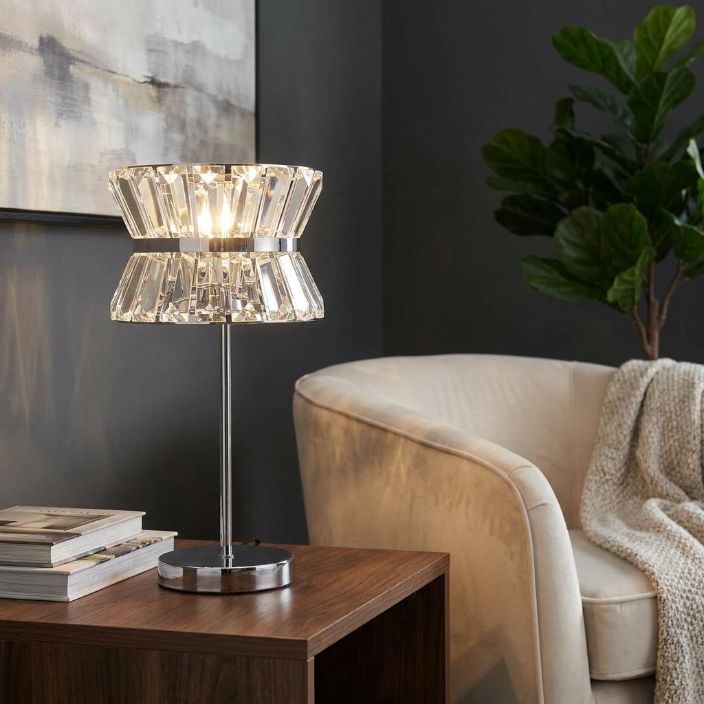 Searchlight Uptown 2 light chrome and crystal table lamp glowing on a wooden table next to a cream armchair.