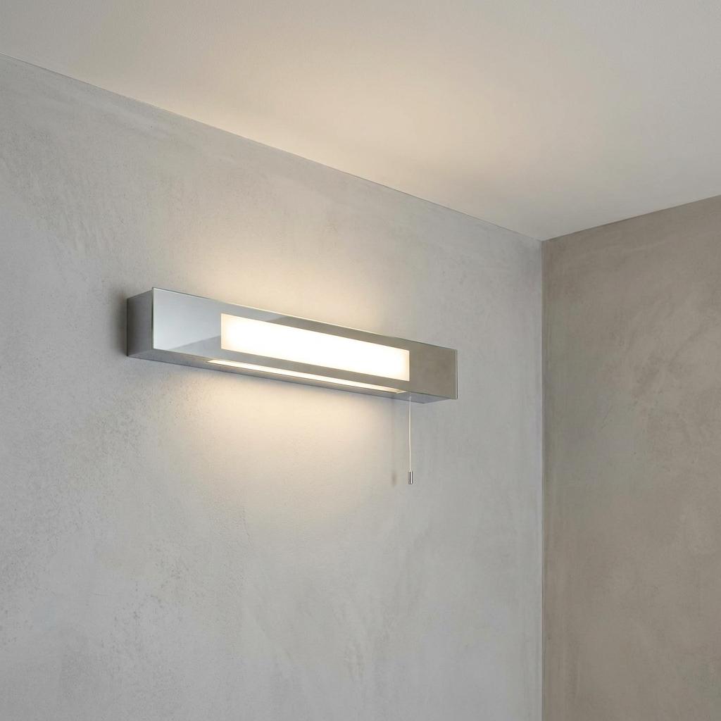 Rectangular chrome shaver light with a frosted glowing diffuser mounted on a gray textured wall with a pull cord.