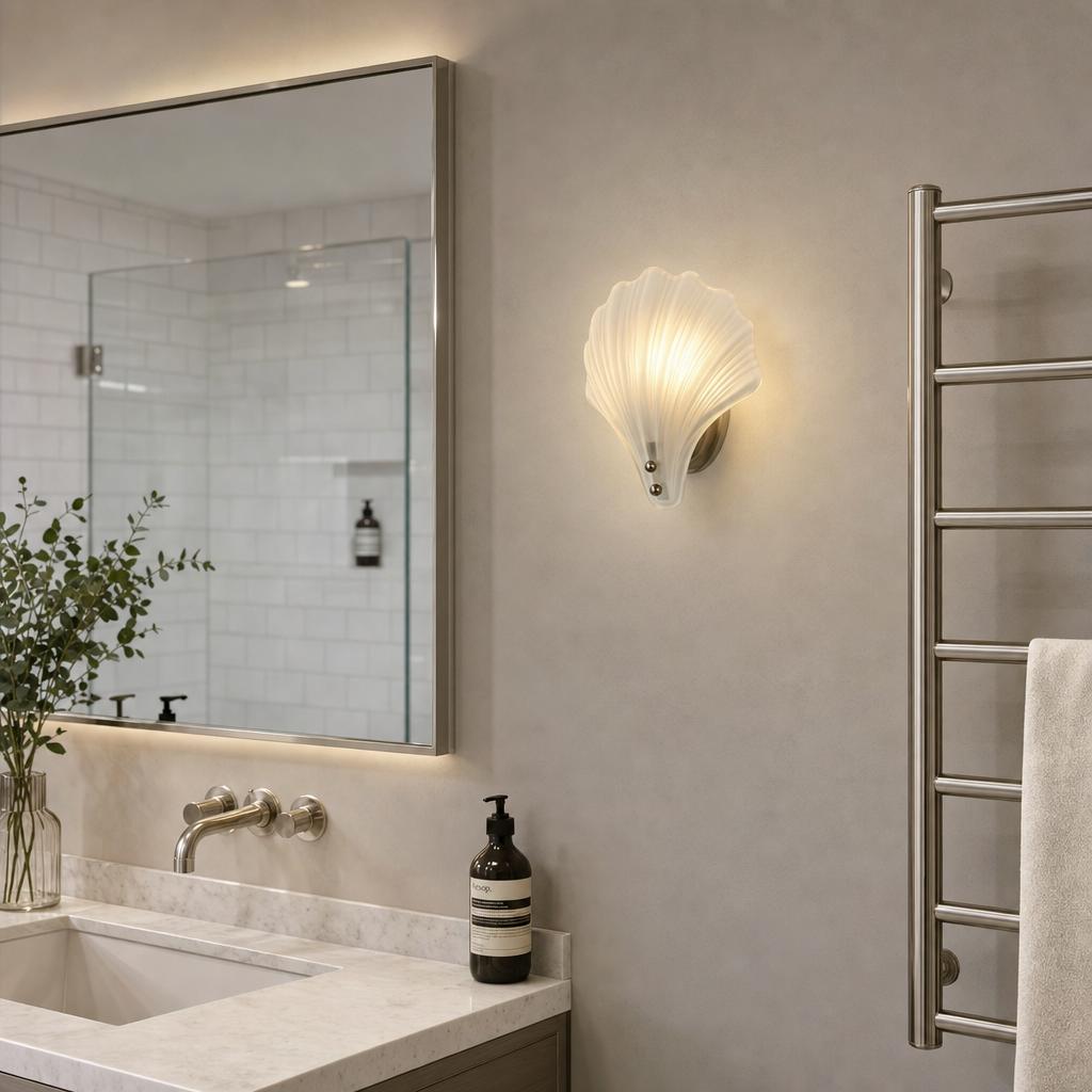 Shell-shaped frosted glass wall light illuminated in a neutral-toned bathroom next to a mirror and towel rack.