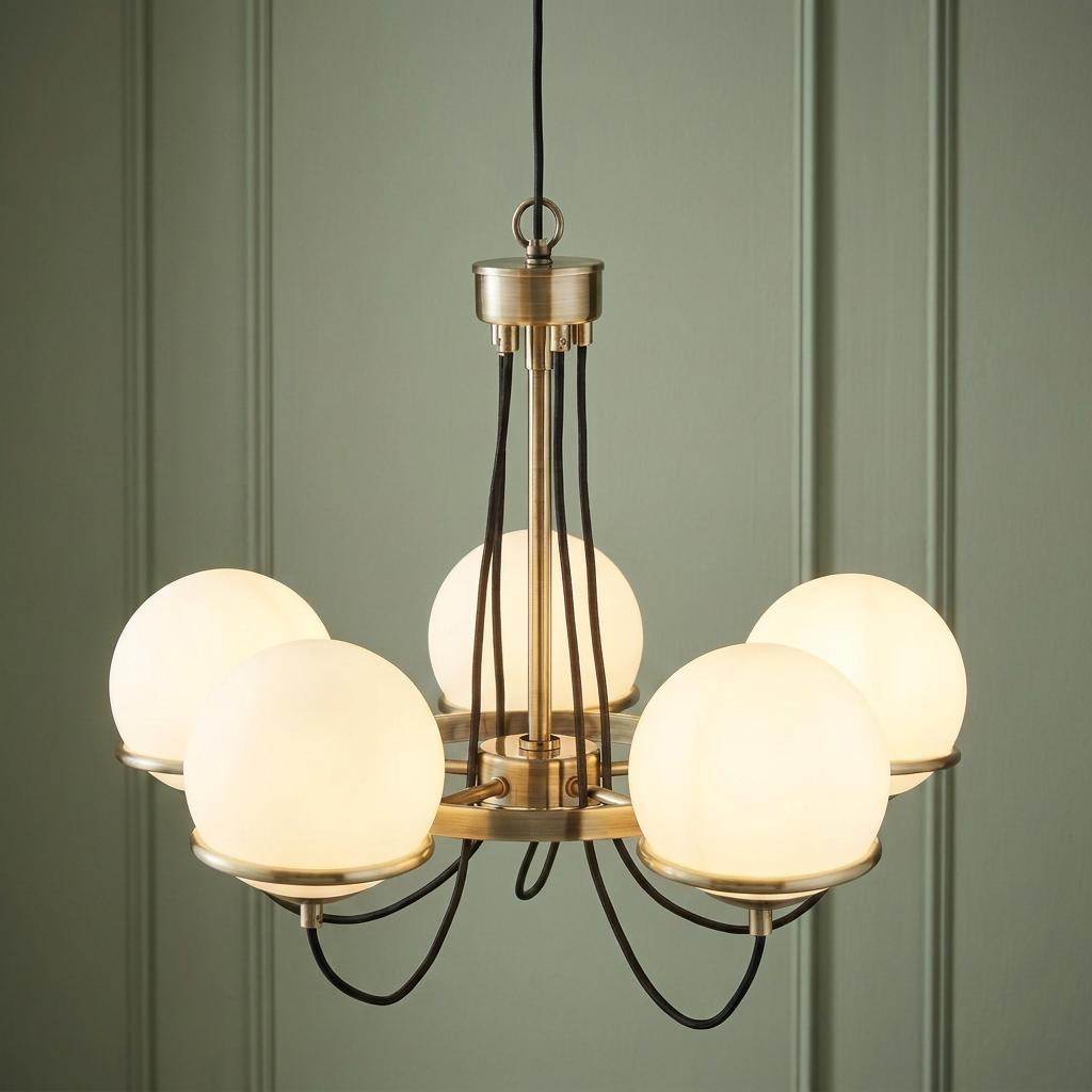 Brass chandelier with five glowing white glass spheres and black braided cables against a paneled green wall.