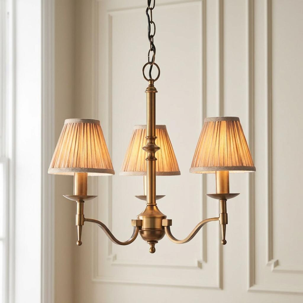 Antique brass 3-light chandelier with pleated beige shades hanging in front of a neutral paneled wall.