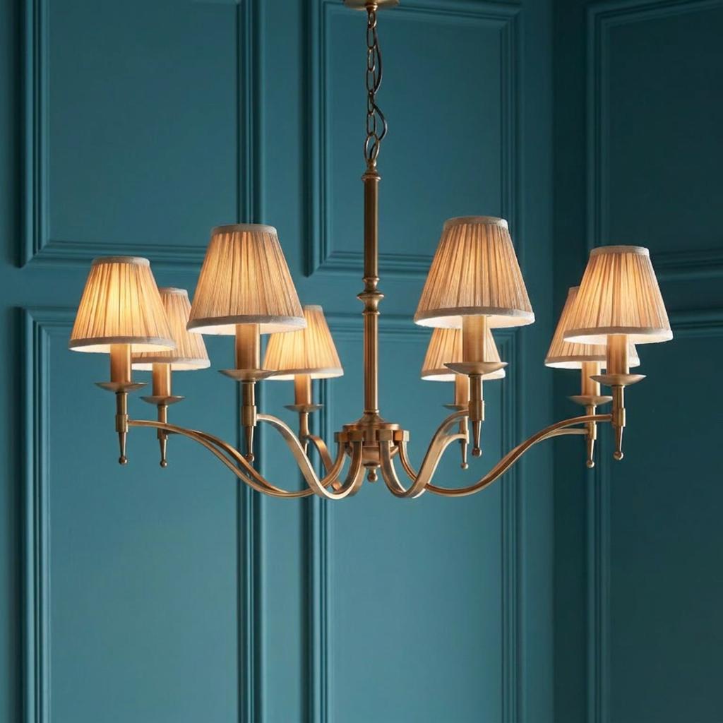 Antique brass 8-light chandelier with beige pleated shades illuminated against a teal paneled wall.