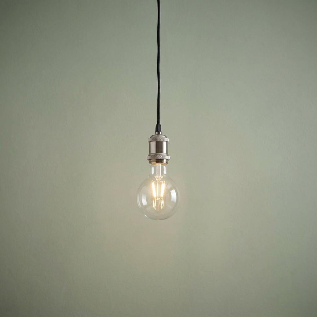 Single lit Edison bulb hanging from a chrome fixture and black cable against a pale sage green wall.