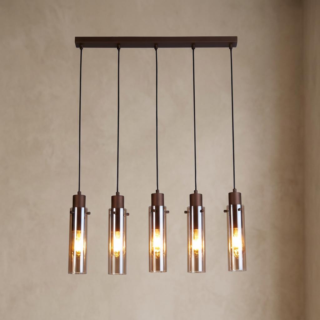 Five amber glass cylinder pendants hang from a mocha metal bar against a beige wall, glowing with warm light.