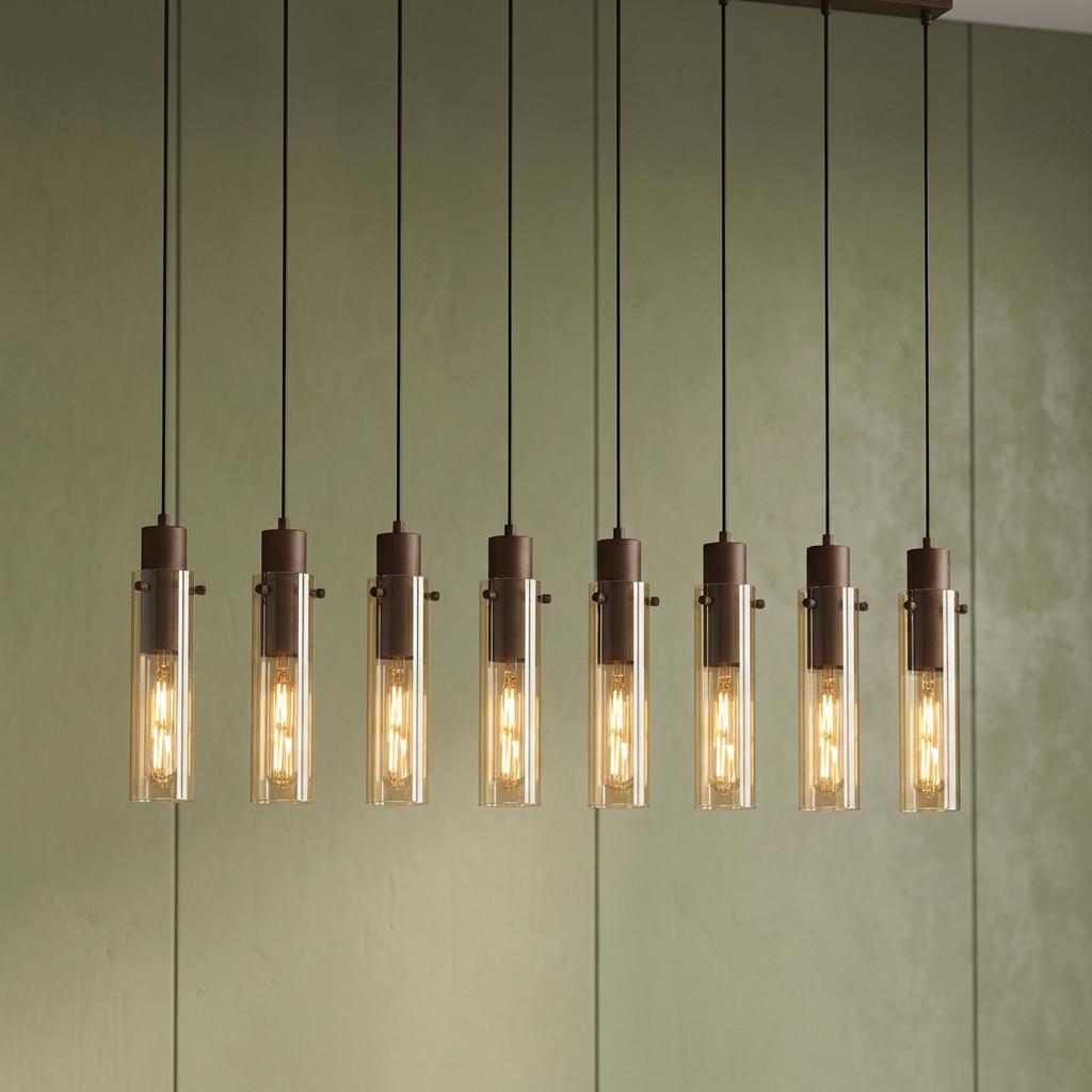 Eight-light mocha bar pendant with cylindrical amber glass shades and glowing bulbs against a green textured wall.