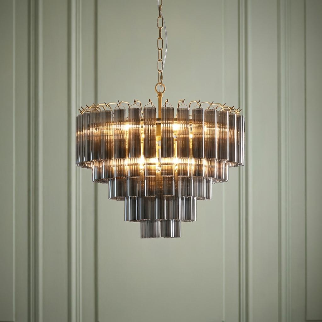 Multi-tiered chandelier with smokey ribbed glass cylinders and brass hardware, lit against a green paneled wall.