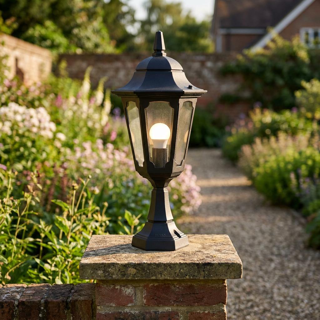 Traditional black outdoor lantern post light illuminated on a brick pillar in a lush garden setting.