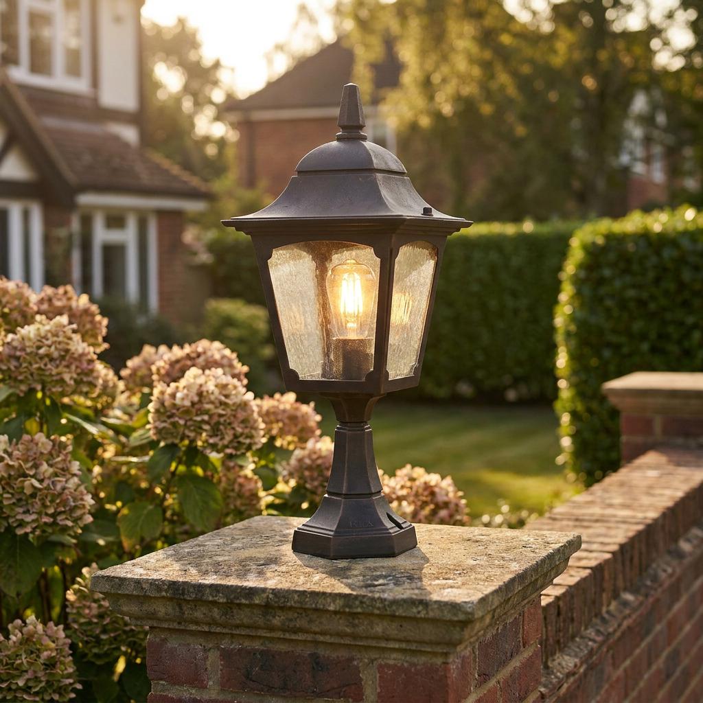 Traditional bronze outdoor pedestal lantern with a warm filament bulb mounted on a stone garden wall pillar.