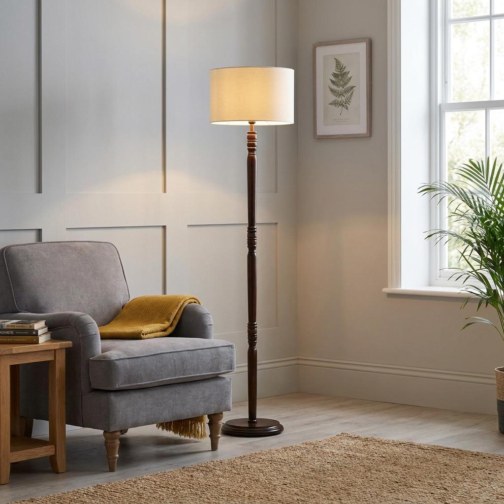 Traditional dark oak floor lamp with a cream shade, lit in a living room beside a grey armchair.