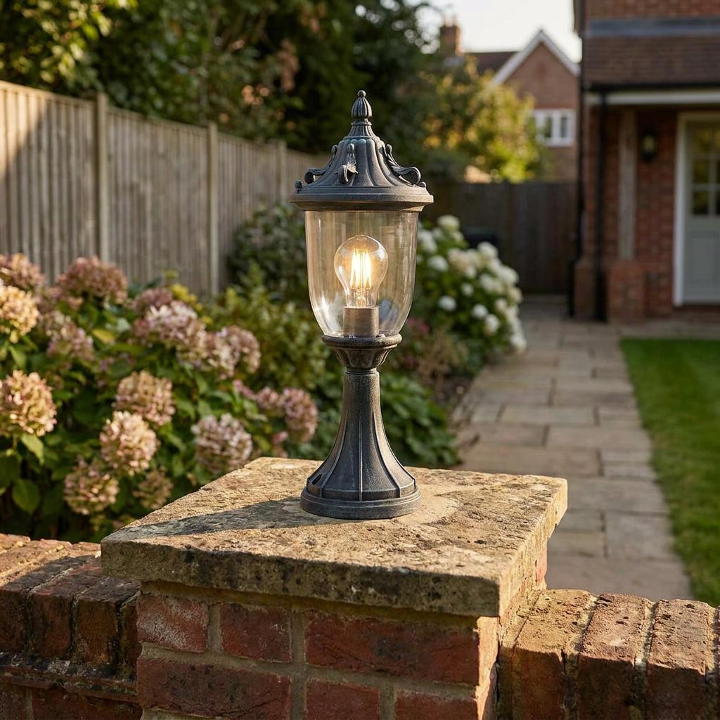 Traditional grey outdoor pedestal lantern with a lit filament bulb, mounted on a stone garden wall pillar.