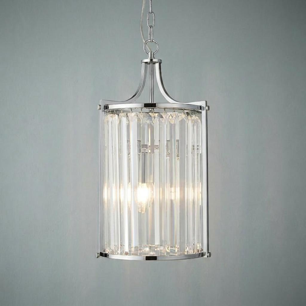 Illuminated chrome and crystal glass cylinder pendant light with vertical prisms against a plain grey wall.