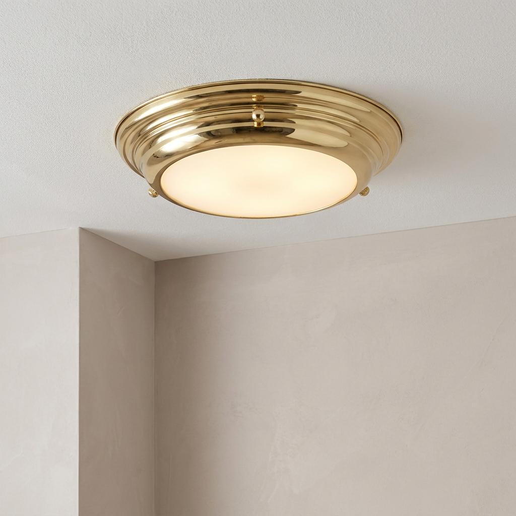 A round polished brass flush mount ceiling light with an illuminated frosted diffuser against a white ceiling.