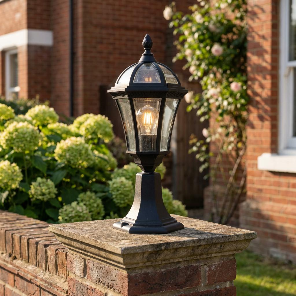 Black lantern-style outdoor post light with a glowing filament bulb mounted on a brick wall pillar in a garden.