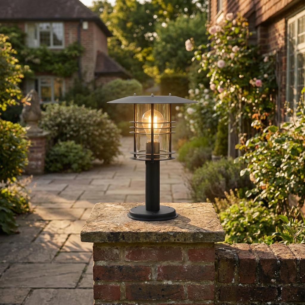 Black outdoor pedestal lantern with a flat top and glowing bulb, mounted on a brick pillar in a lush garden.