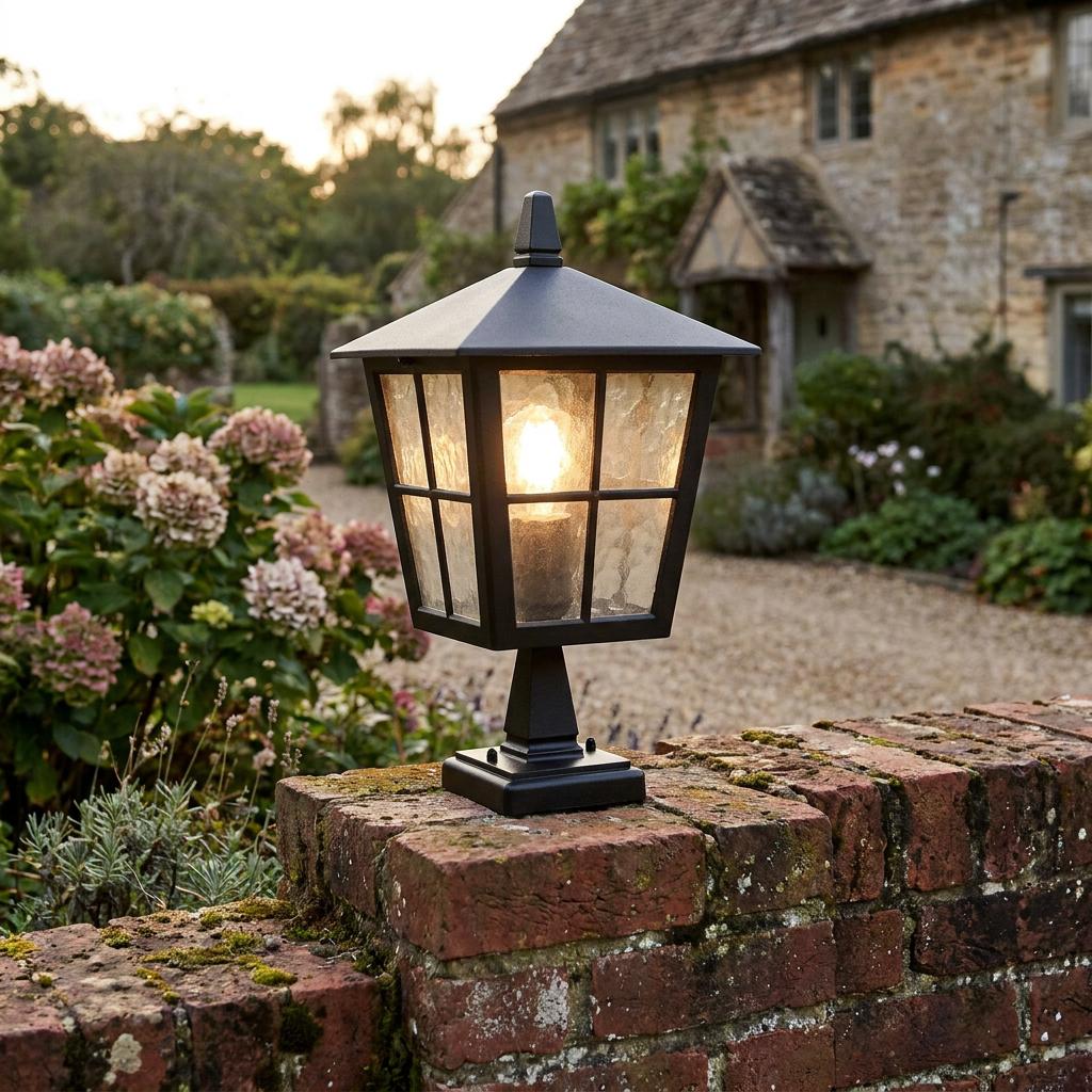 Black traditional lantern pedestal light glowing on a rustic brick wall in a lush garden setting.