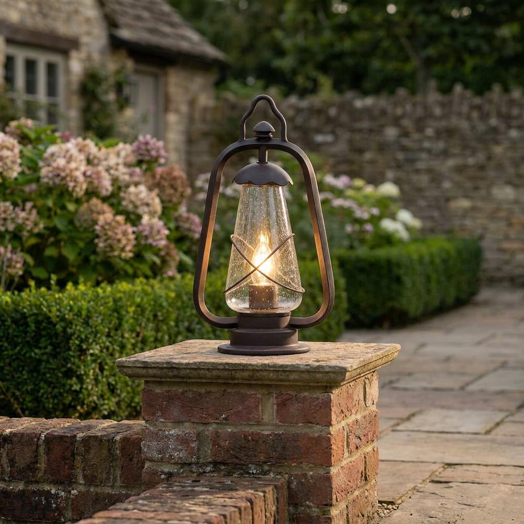 Bronze lantern-style outdoor light with seeded glass and a glowing bulb sits on a stone pillar in a garden.