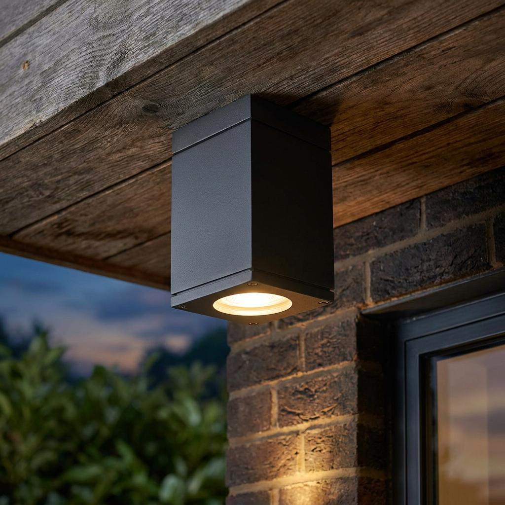 Modern black cube outdoor light fixed to a wooden ceiling, illuminating a brick wall at dusk.