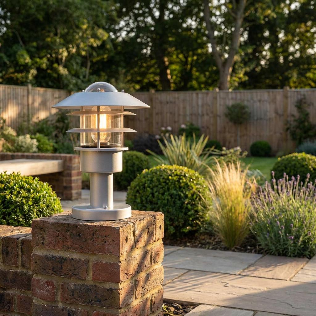 A silver tiered pedestal light glowing on a brick pillar in a lush garden setting.