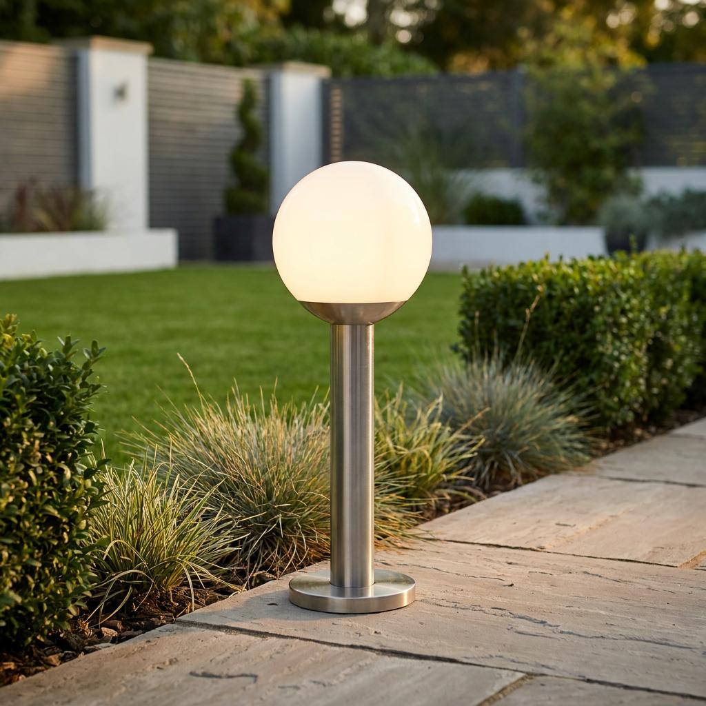 A glowing white globe light on a stainless steel pedestal base situated on a stone garden path.