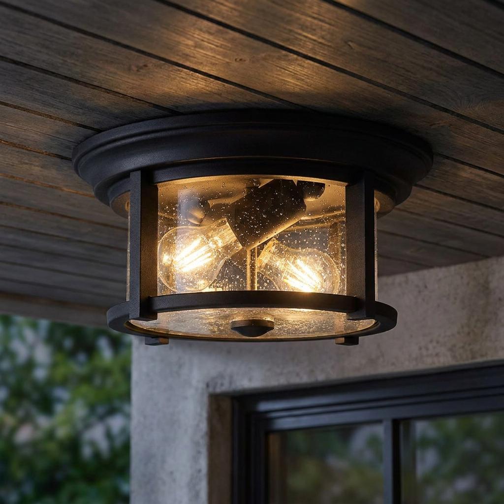 Outdoor flush mount ceiling light with a black metal frame and seeded glass housing two glowing bulbs on a wood porch.