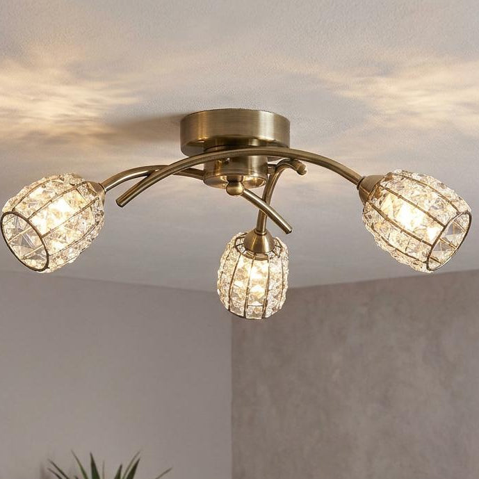 A three-light flush mount ceiling fixture with a brushed gold/brass finish and clear crystal-encrusted globe shades, shown illuminated in a modern living room setting.