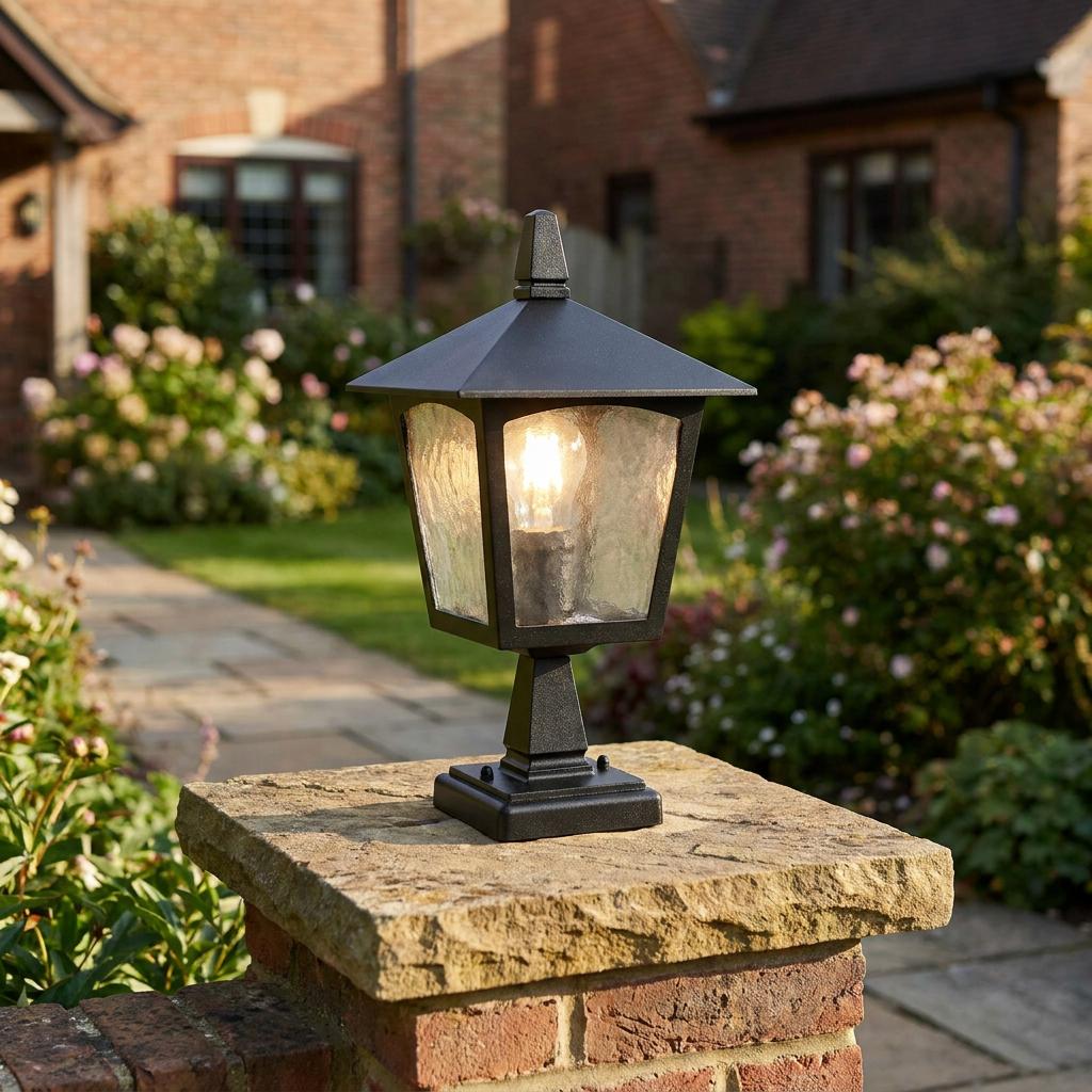 A black outdoor lantern with rippled glass panels glows on a stone wall pillar in a garden.