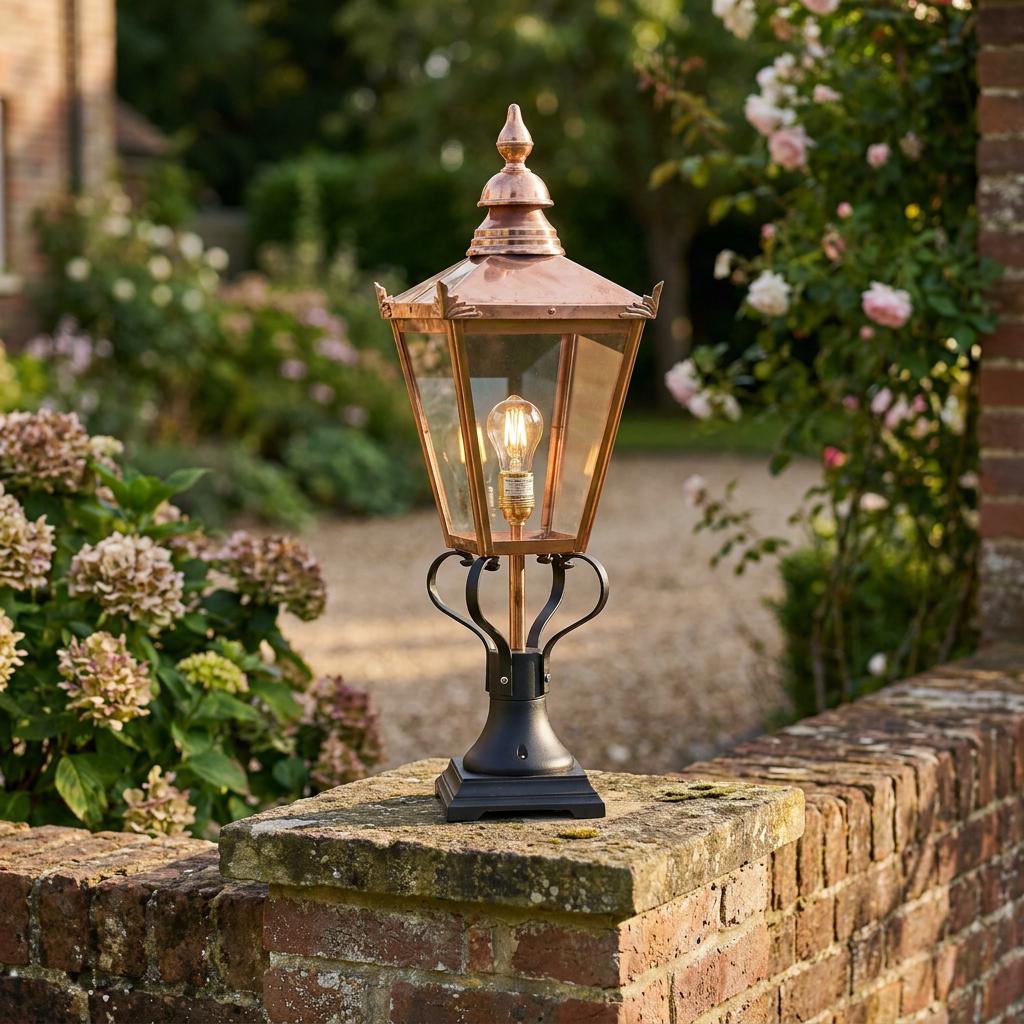 Traditional copper lantern with a glass housing and black scrolled base, mounted on a brick wall in a blooming garden.
