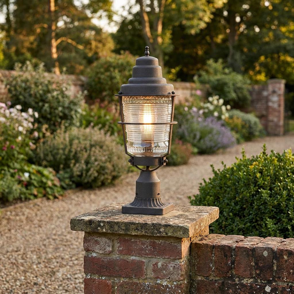 Traditional dark grey pedestal lantern with ribbed glass glowing on a brick pillar in a lush garden.